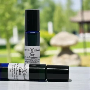 Blue roll-on bottle labeled Sacred Blend Joy essential oil, 10ml, with a black cap, set against a blurred outdoor background featuring greenery and a stone structure.