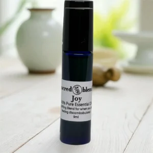 Joy 10ml roll-on essential oil bottle from Sacred Blends in a dark blue glass with black cap on a light wooden surface; label shows Joy and 100% Pure Essential Oil.