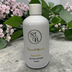 A Nourish 250ml bottle of Sacred Blends Day Face and Body Crème stands among green leaves and pink flowers.